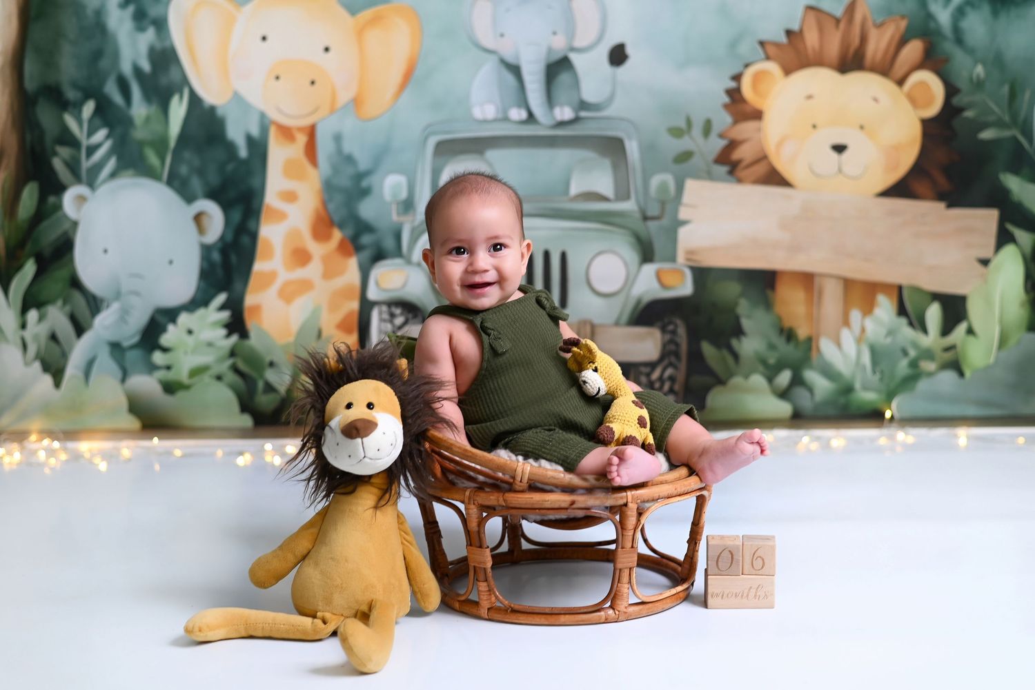 A baby sits on the floor with stuffed animal toys against a whimsical forest-themed wall mural.