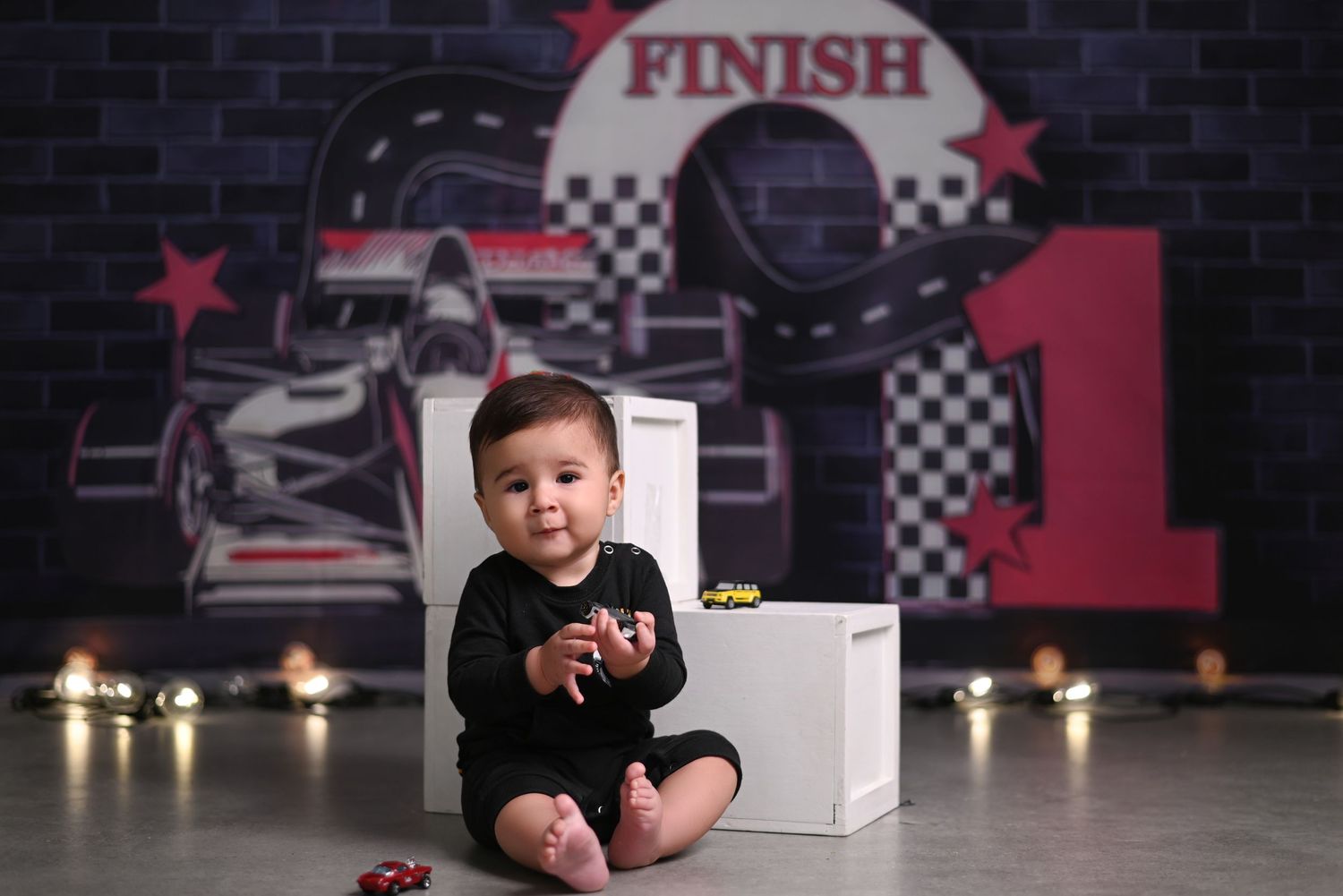 Racing themed first birthday photo shoot with baby in black outfit posing with cake and decorations.