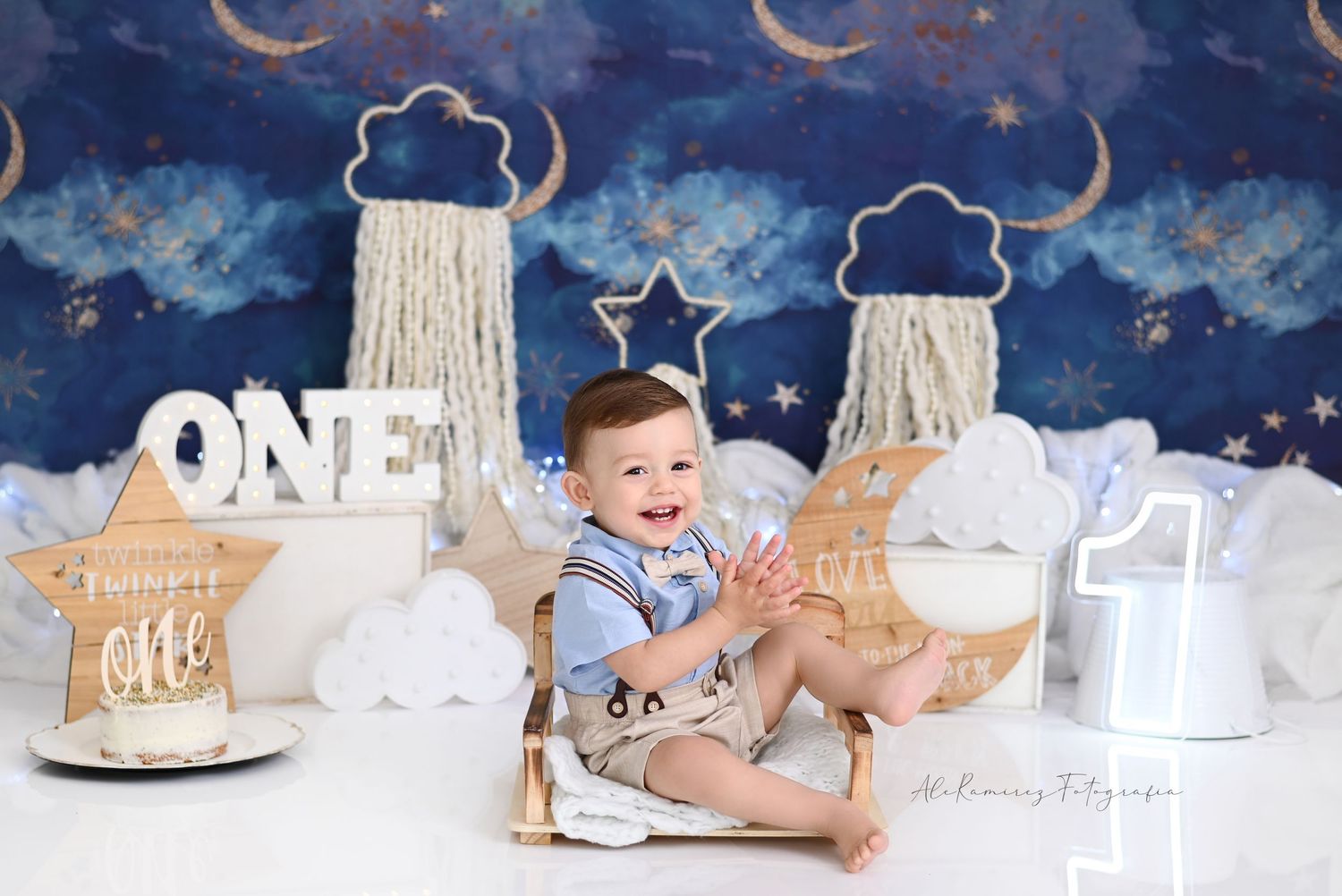 Baby in blue shirt sits on cloud backdrop with ethereal nighttime decor for first birthday photo session.
