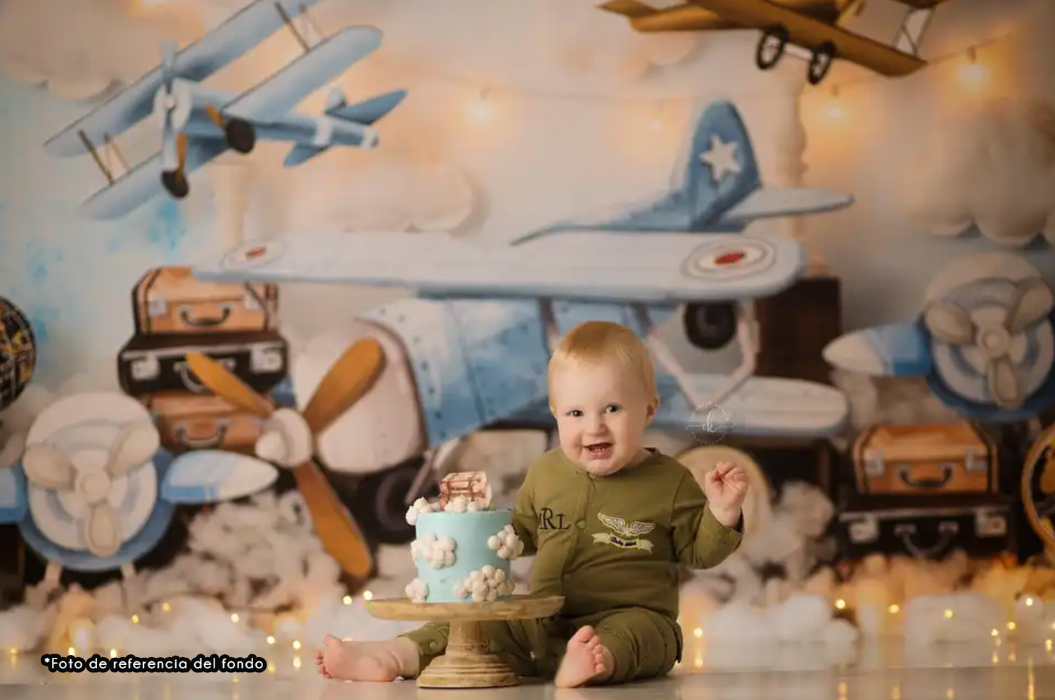Baby in green onesie sits on floor against vintage aviation themed backdrop with toy planes and suitcases.