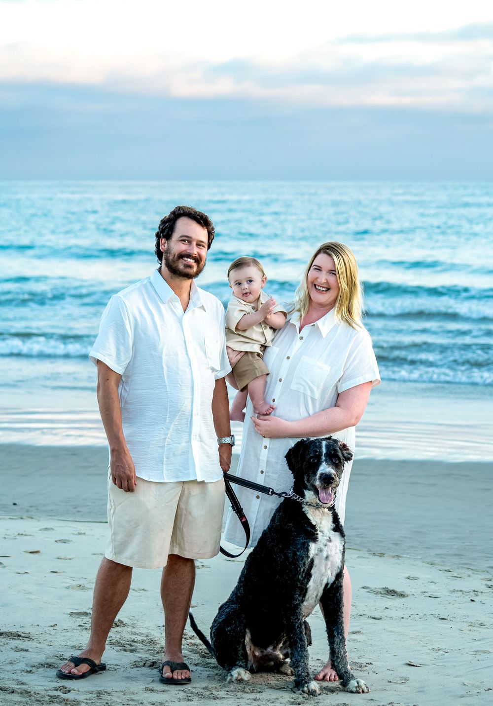 Family in white outfits posing together with their dog on beach at sunset.
