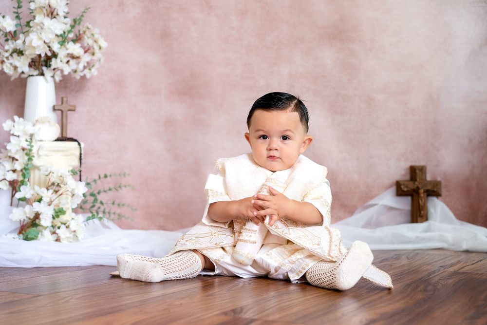 Baby boy in a cream embroidered christening outfit sits on a wood floor beside small wooden crosses and white flower arrangements.
