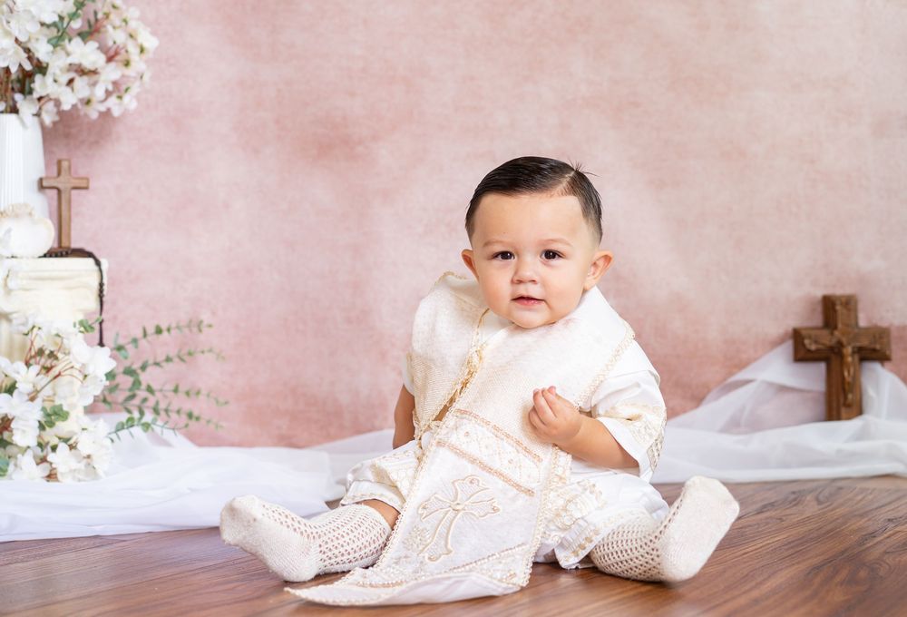 Baby boy in an ivory embroidered christening outfit and white booties sits on a wood floor beside wooden crosses and white flowers.
