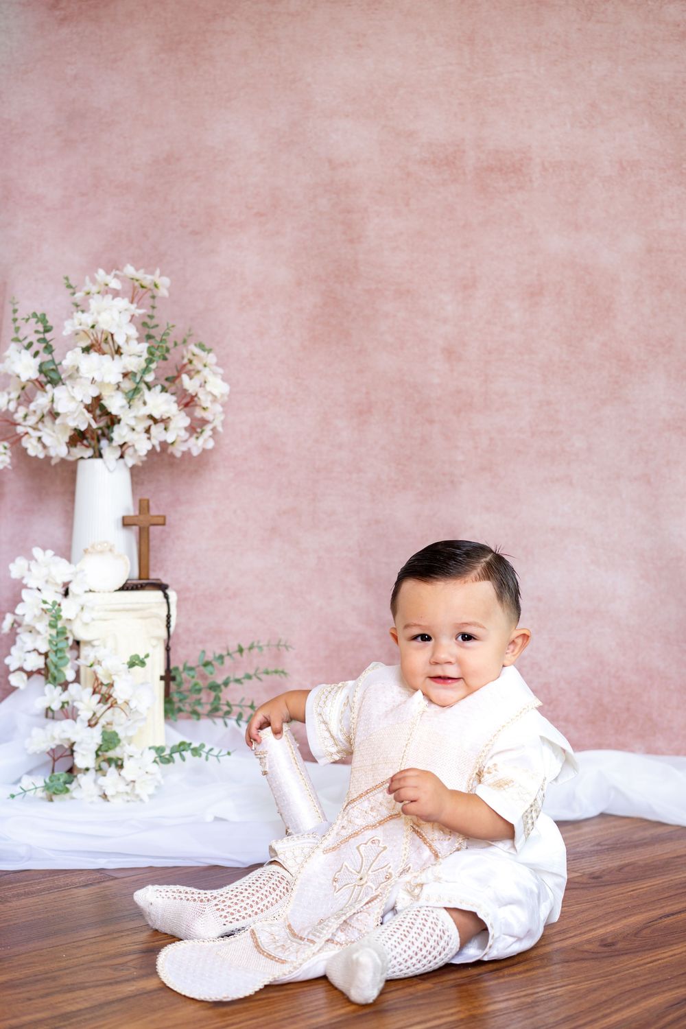 Smiling baby boy in a cream embroidered baptism outfit sits on a wood floor holding a candle beside white flowers and a small wooden cross.