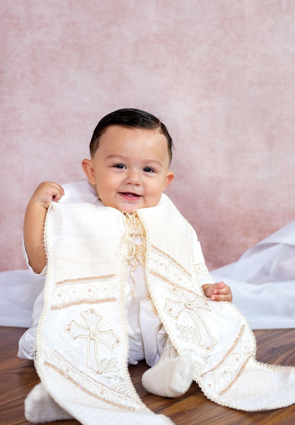 Smiling baby boy in a white christening outfit with an embroidered cream stole featuring gold crosses, seated against a soft pink backdrop.