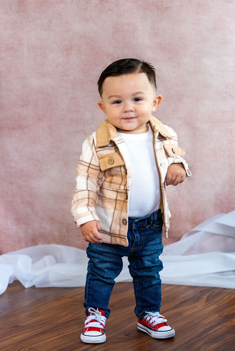 Smiling toddler boy in a plaid jacket, white shirt, jeans and red sneakers standing on a wood floor against a pink backdrop.