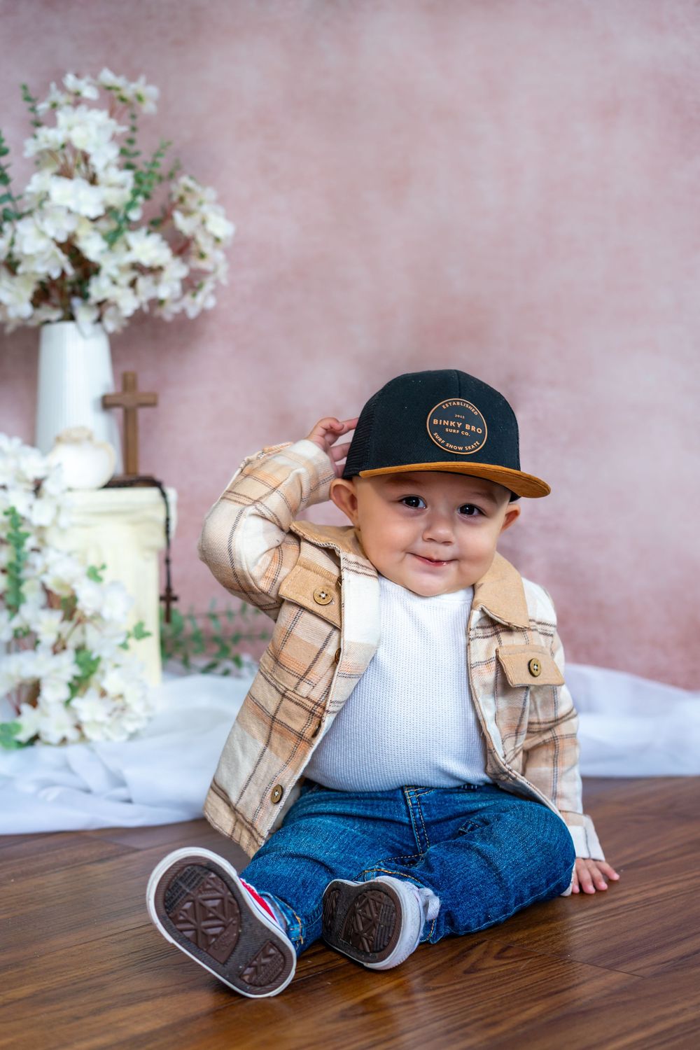 Happy baby boy in a plaid jacket and black cap sitting on a wood floor beside white flowers and a small cross in a portrait.