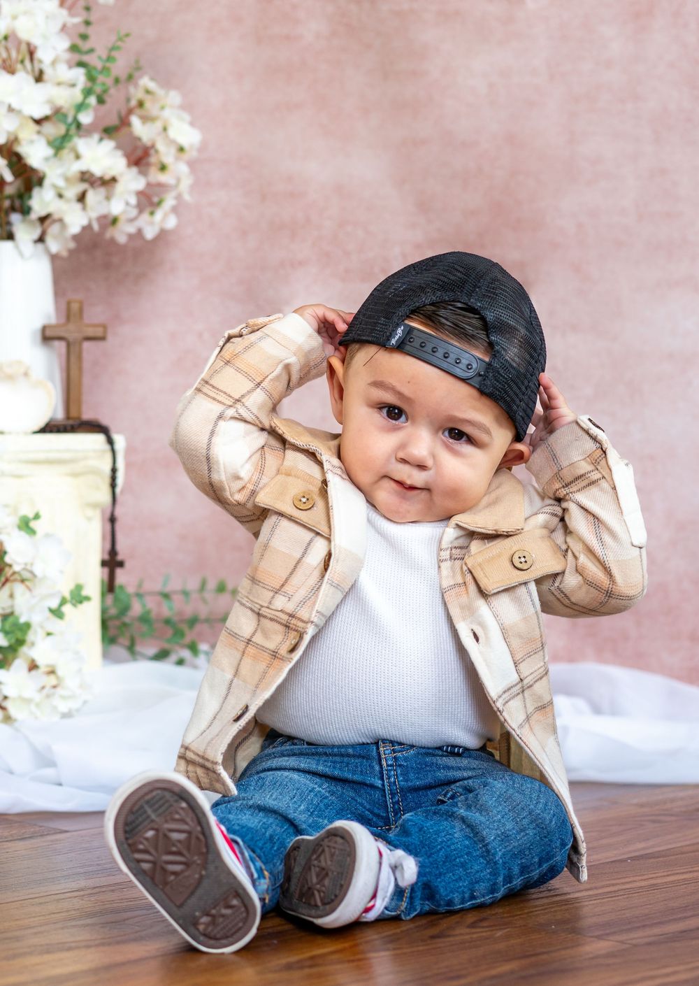 Toddler in a backward black cap, plaid jacket and jeans sits on a wood floor holding his cap, beside white flowers and a wooden cross.
