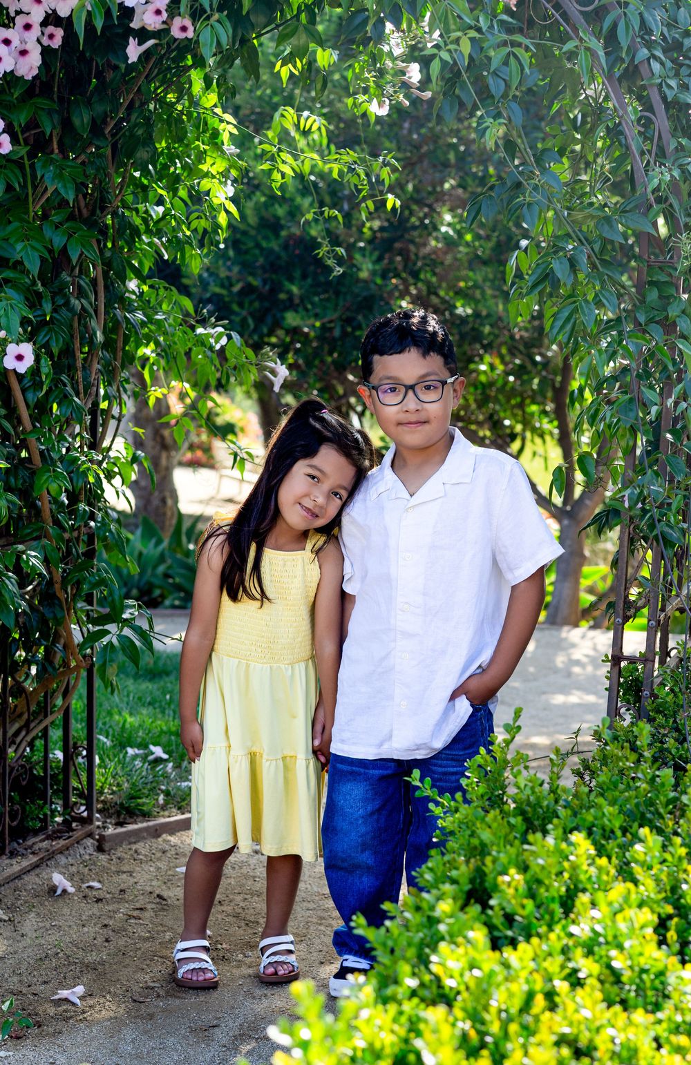 Two children pose in a lush garden, girl in yellow dress and boy in white shirt with glasses.