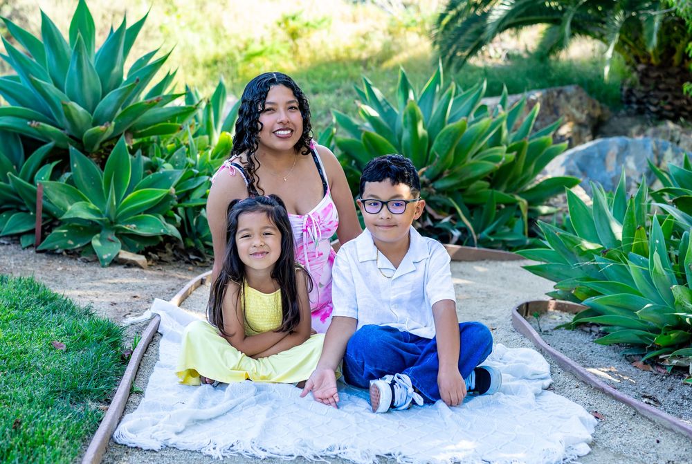 A woman and two children smile together on a white blanket surrounded by lush green agave plants outdoors.