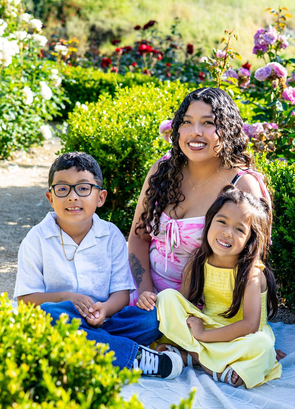 A smiling woman with two young children sitting outdoors in a lush garden with colorful roses blooming in the background.