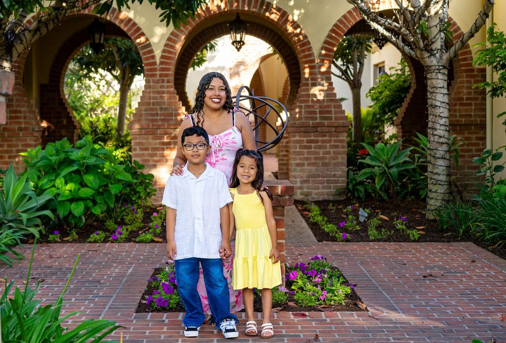 A woman poses with two children in front of elegant brick arches and lush greenery at a scenic outdoor venue.