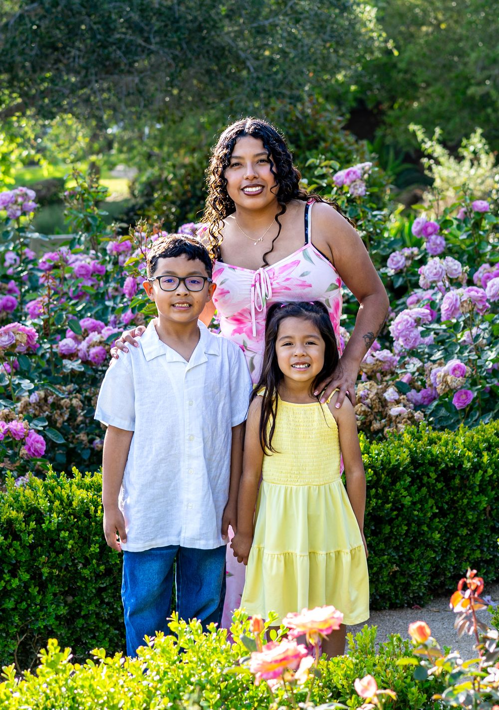A woman and two children smile together in a lush garden filled with pink roses and greenery.