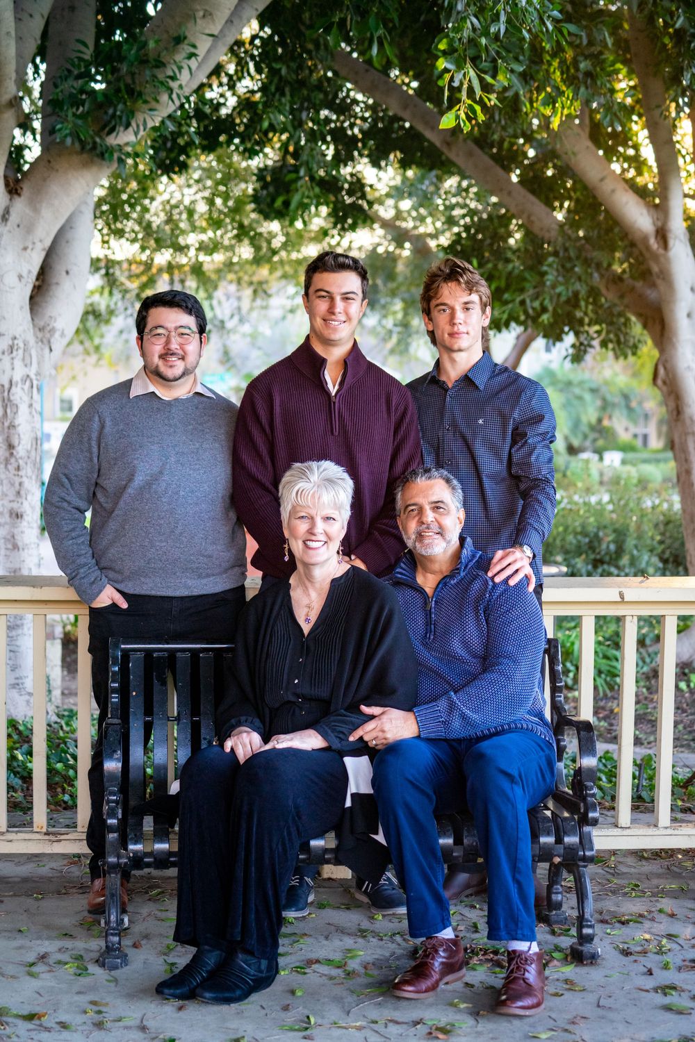 A family group sits together on a wooden porch bench surrounded by leafy trees on a sunny day.