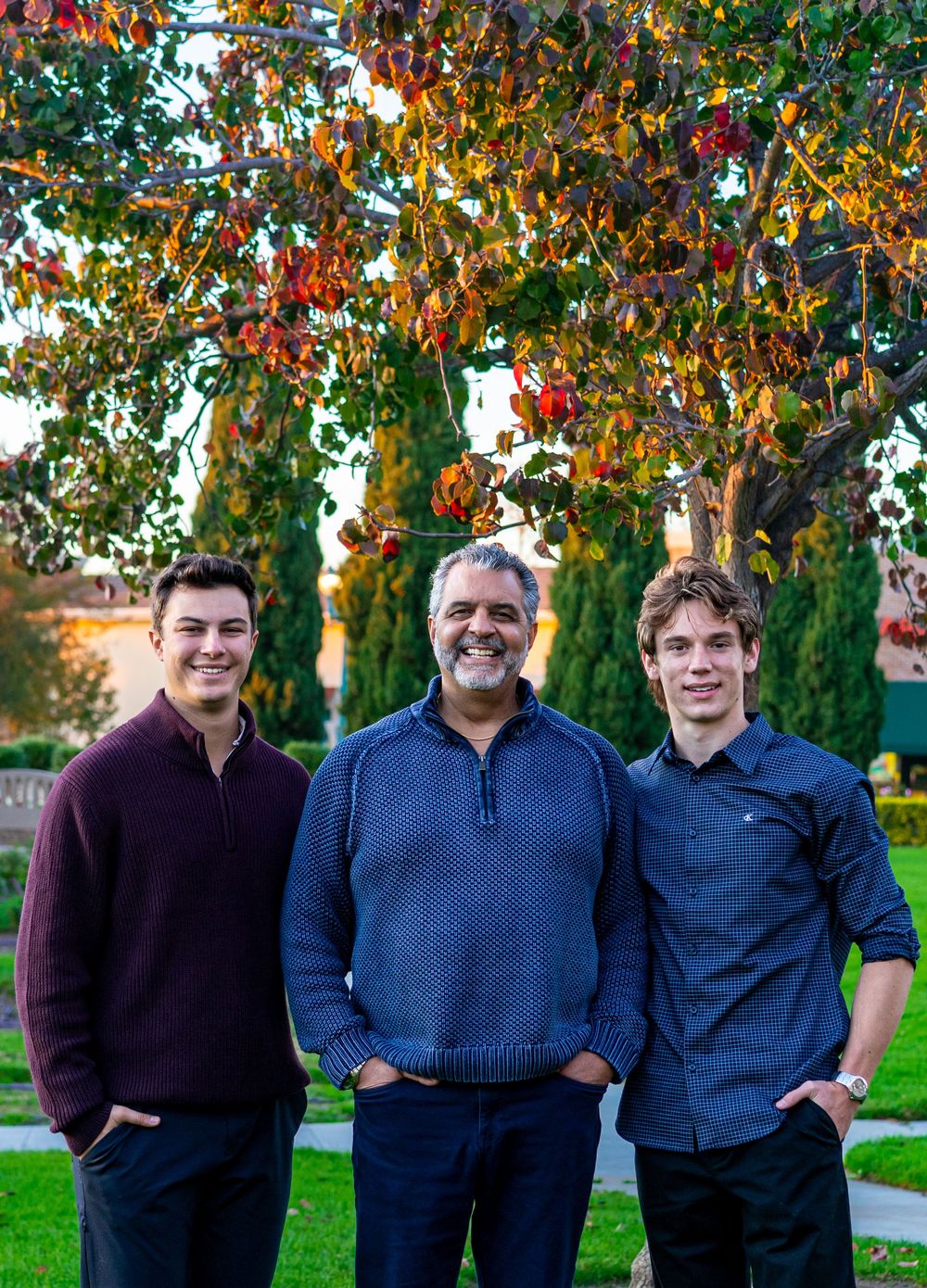 Three people stand together outdoors in front of autumn trees wearing dark colored shirts.