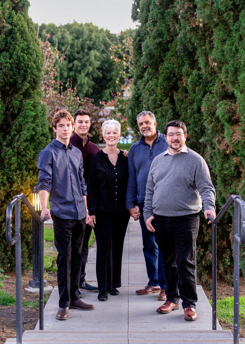 A group of five people standing together on a garden path lined with tall evergreen trees and flowering bushes.