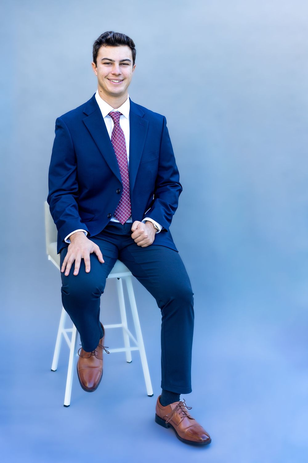 Professional business portrait of someone in a navy suit and pink tie seated on a white stool against a blue backdrop.