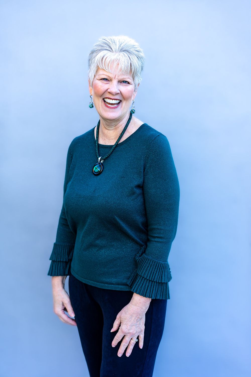 A person wearing a black sweater with ruffle sleeves and black pants poses against a light blue background.