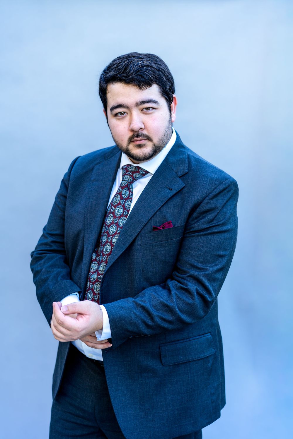 Professional portrait of a person in a navy suit with floral tie against a light blue background.
