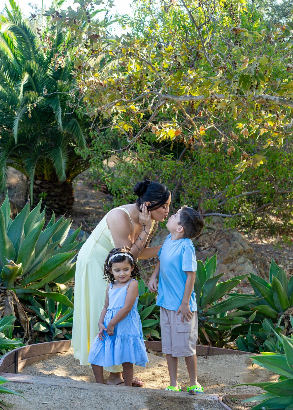 A mother in a yellow dress kisses her young son while her daughter stands beside them in a lush garden.