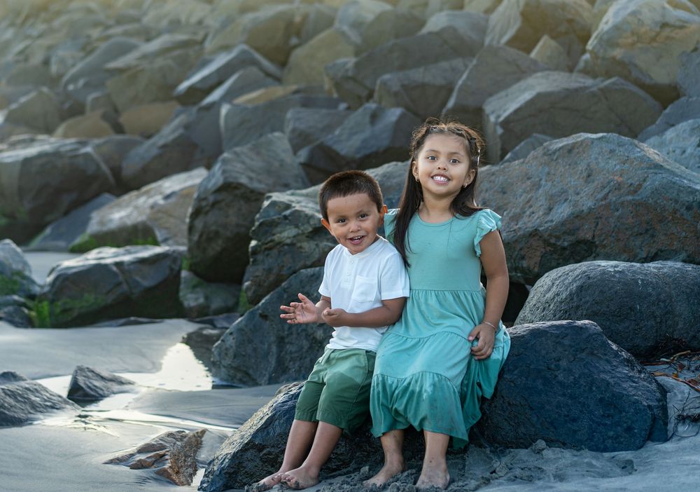 Two young children smile on rocky beach, boy in white shirt and green pants, girl in teal dress.