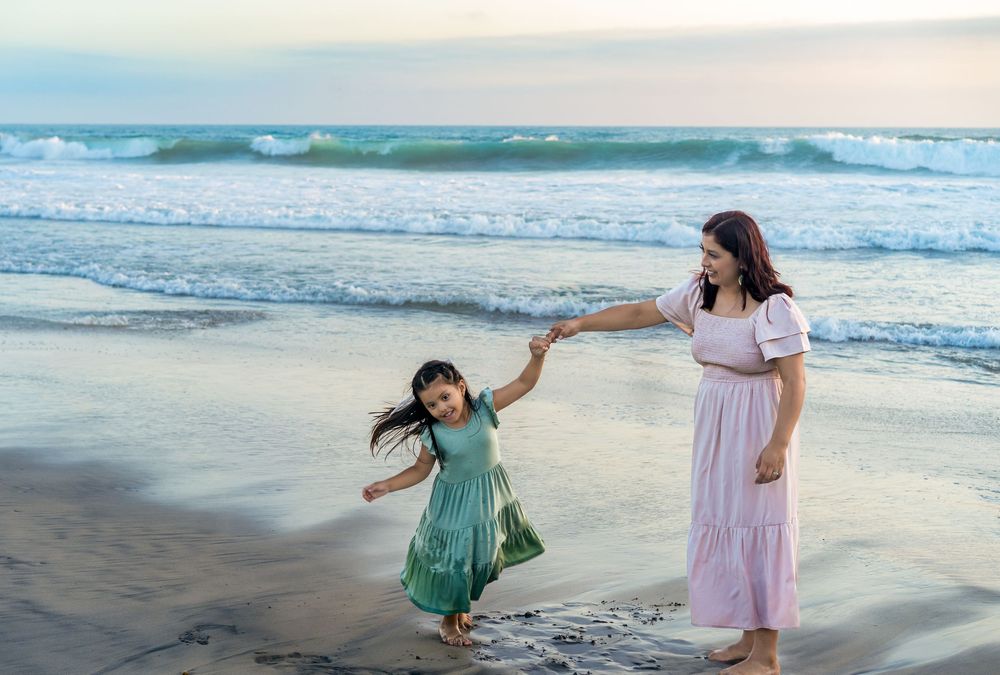 A mother and young daughter dance joyfully on a sandy Carlsbad beach at sunset, wearing flowing dresses by ocean waves.