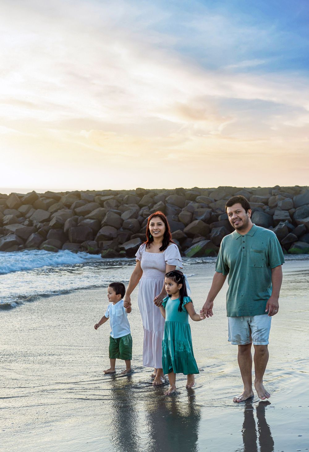 Family of four walking on Carlsbad beach at sunset, with rocky shoreline and waves in the background.