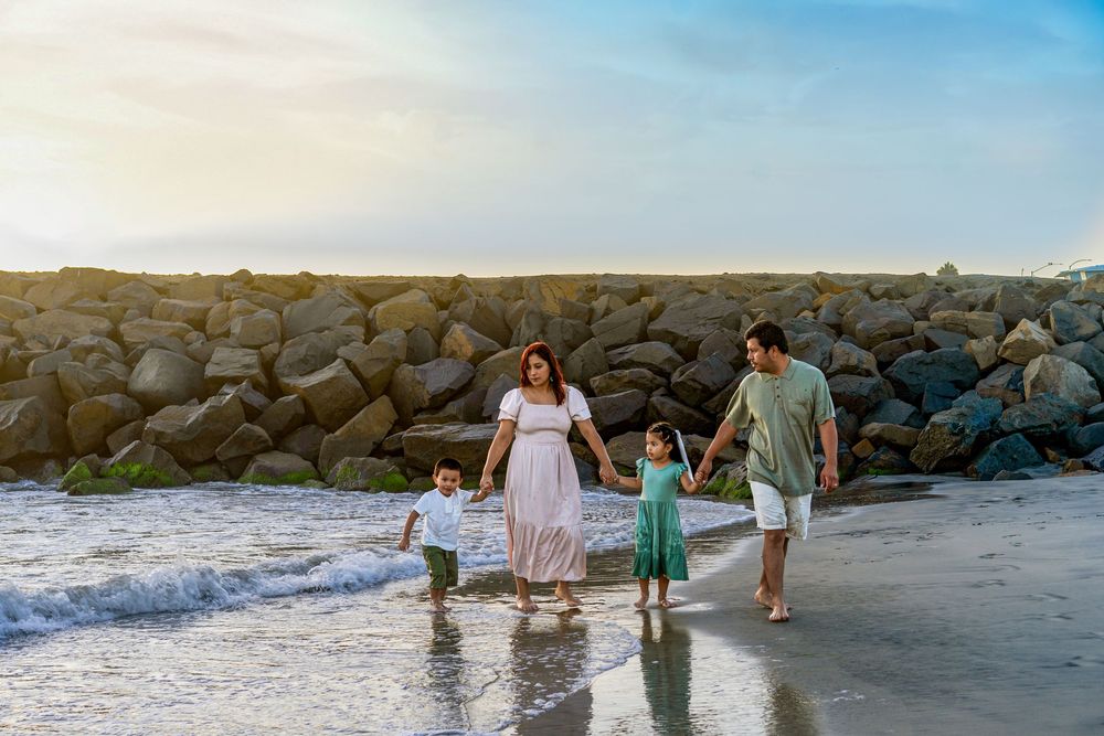 Family of four walking along a sandy beach near a rocky jetty at sunset, with waves gently lapping at their feet.