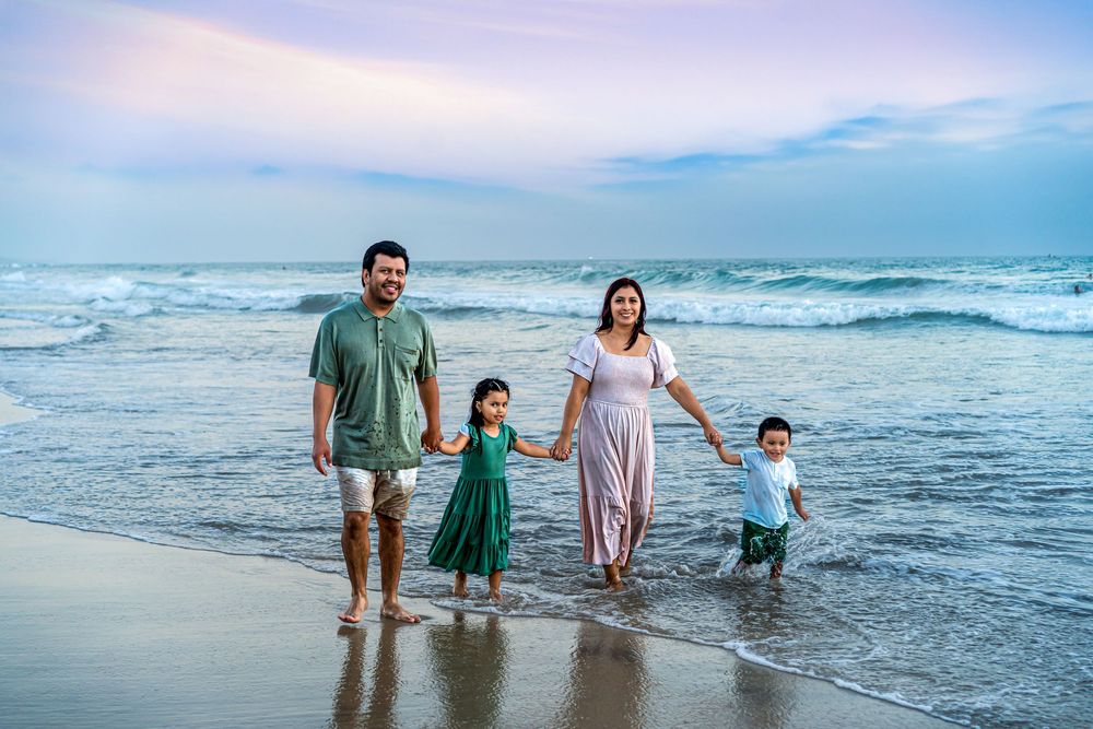 Family of four walking hand-in-hand on a beach at sunset, waves crashing behind them.