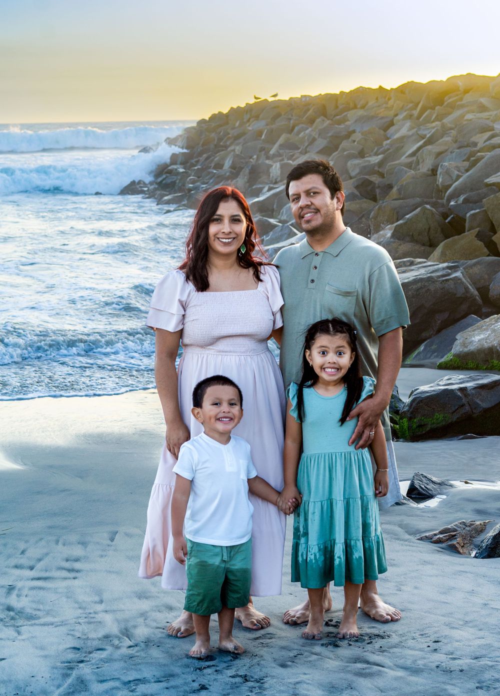 Family of four smiling together on a sandy beach at sunset near rocky jetty.