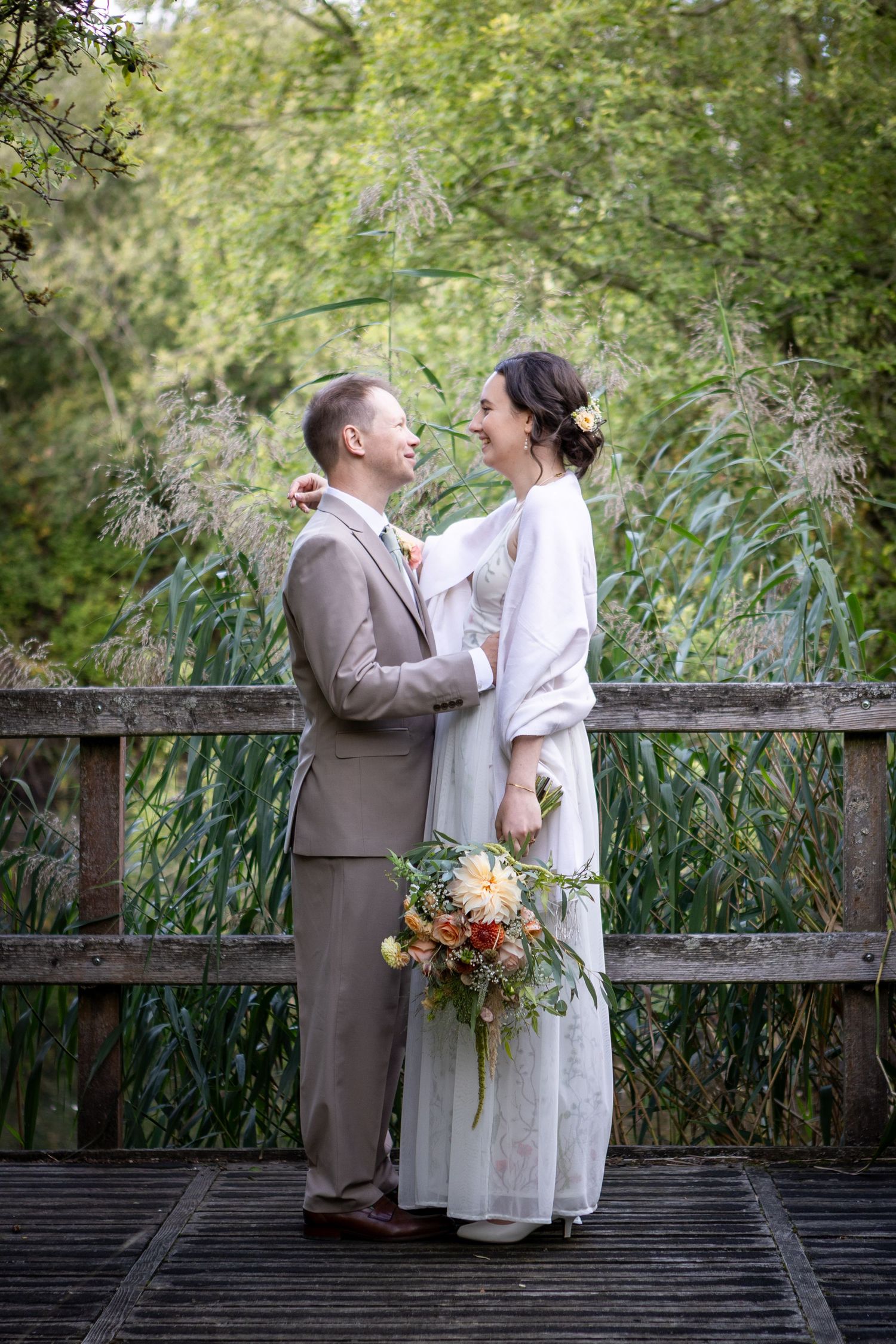 Bride and groom laugh together on wooden bridge surrounded by tall reeds, she holds colorful bouquet.