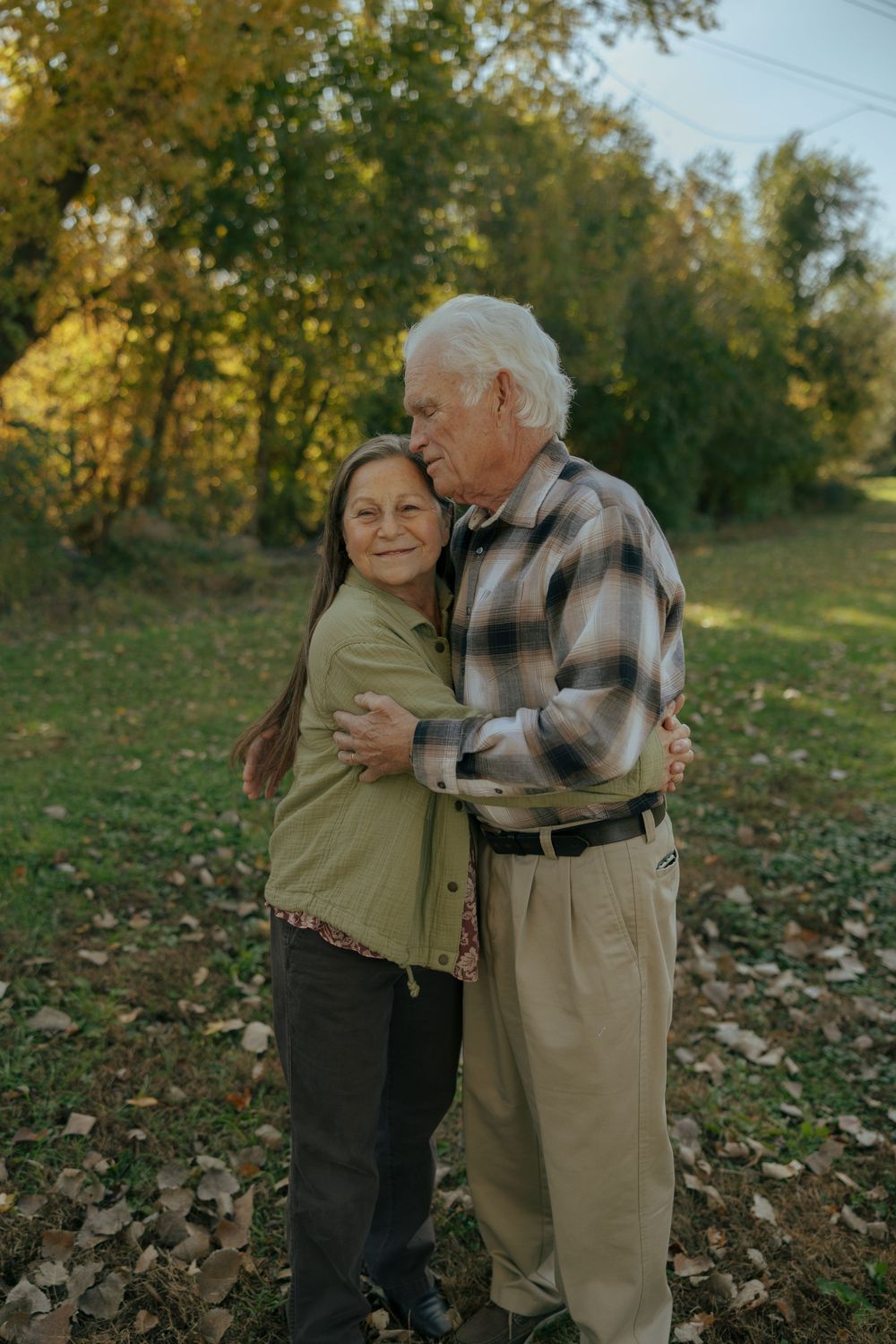 Elderly couple embraces tenderly in an autumn park with fallen leaves on the ground during golden hour.