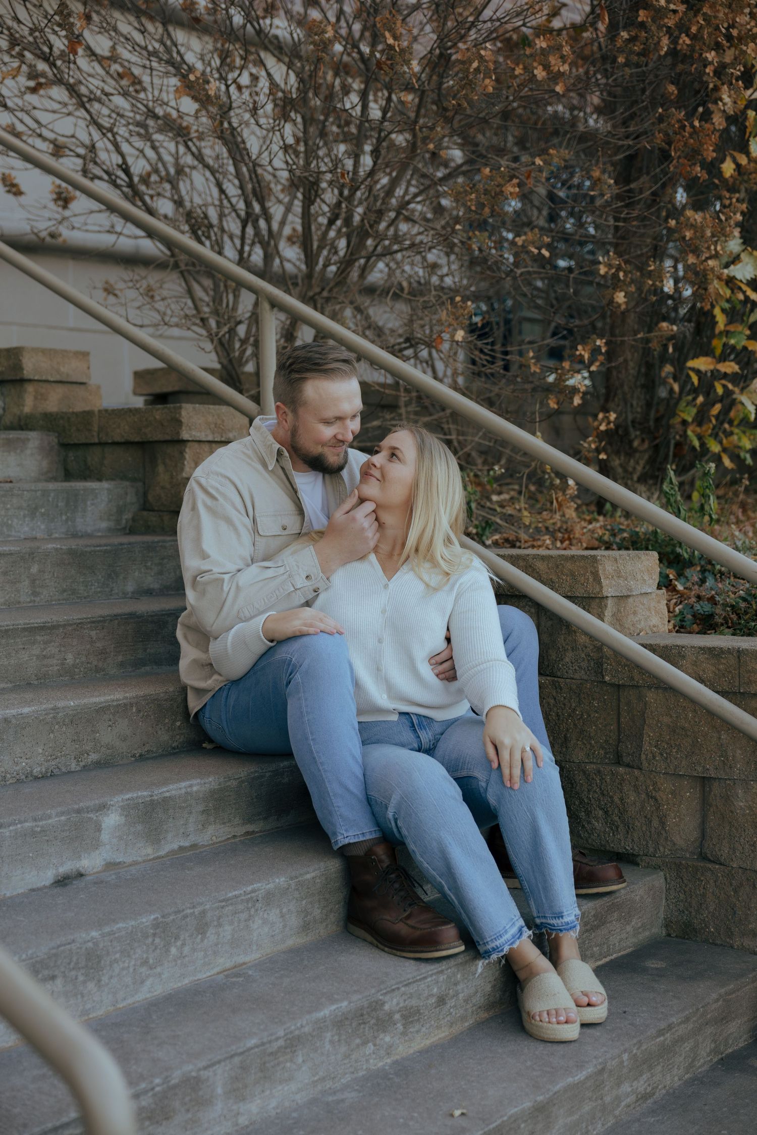 A couple sits together on stone steps outdoors during autumn, sharing an intimate moment in casual attire.