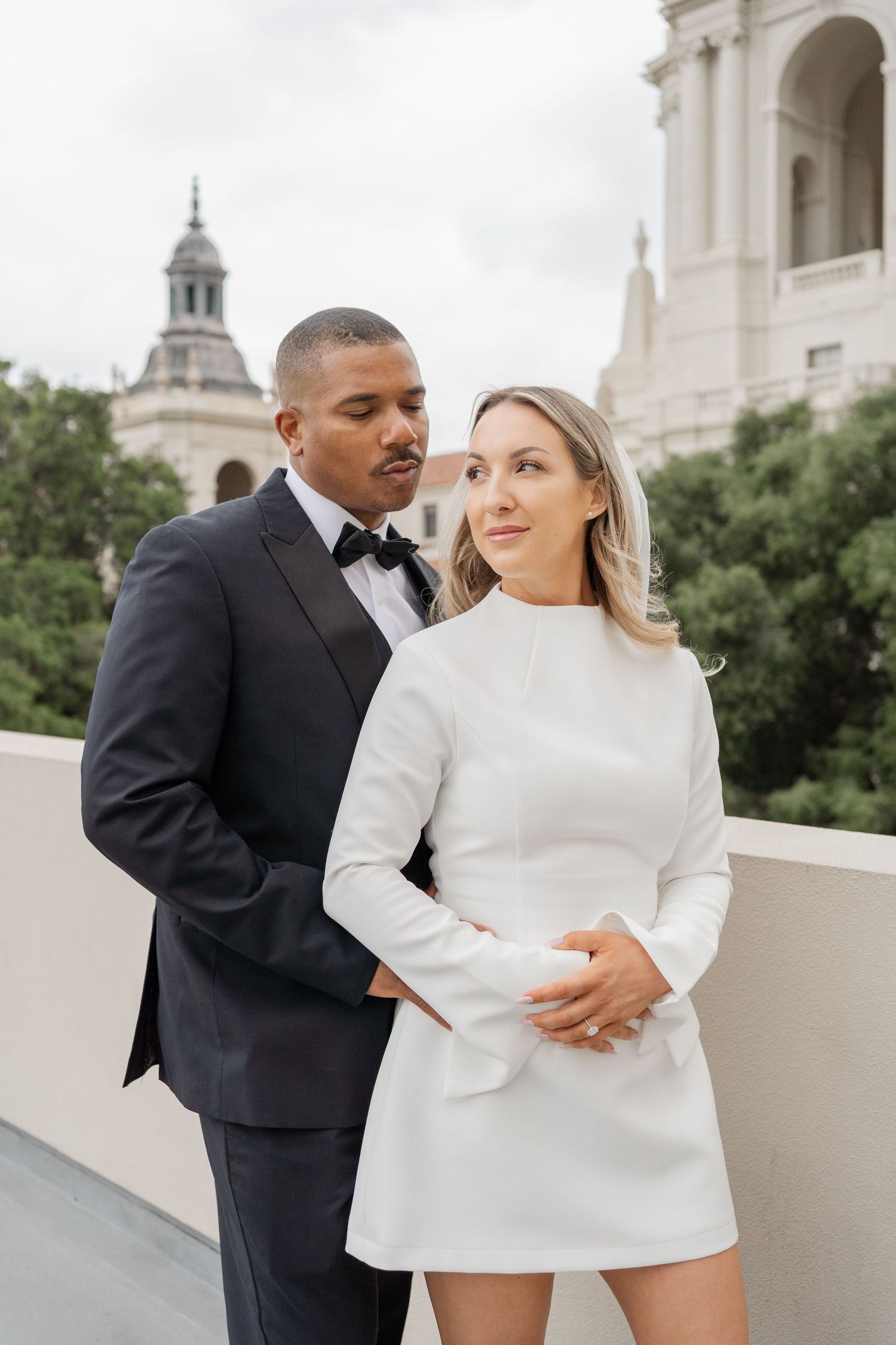 Elegant couple in formal attire poses on rooftop with domed building in background.