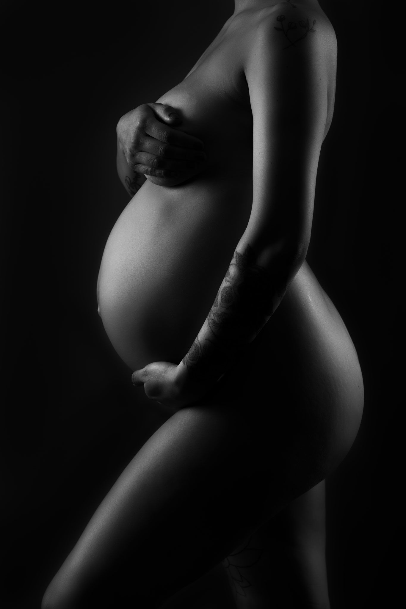 Artistic black and white side view silhouette of pregnant belly and torso against dark background.