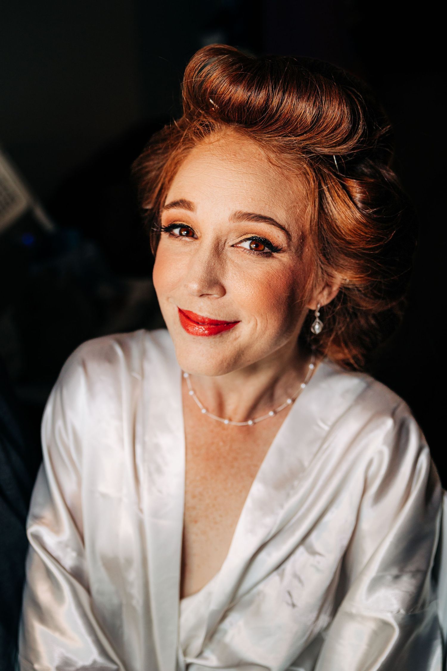 A portrait of a bride with red hair wearing a white silk robe and delicate pearl necklace smiling warmly at the camera.