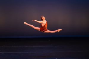 A dancer in brown attire performs a dramatic split leap against a deep blue background on stage.