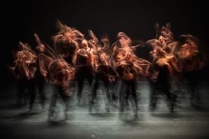 Multiple dancers create a dynamic motion blur effect in brown tones against a dark background.