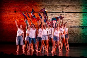 Dancers in white costumes perform a synchronized routine against a red and green lit brick wall.