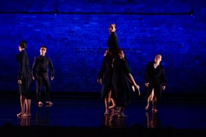 Silhouetted dancers perform against a blue-lit brick wall in a dramatic contemporary piece.