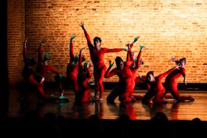Dancers in red costumes perform dynamic movements against a brick wall backdrop.