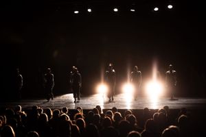 Stage performers silhouetted against bright backlighting with audience visible in foreground.