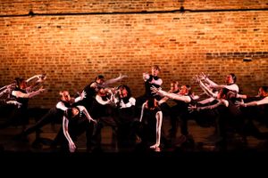 Dance ensemble performs synchronized movements against rustic brick wall.