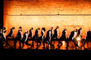 Dancers in silhouette moving in line formation against warm-lit brick wall background.