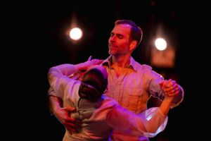 Two dancers perform an intimate ballroom dance under dramatic purple lighting on a dark stage.