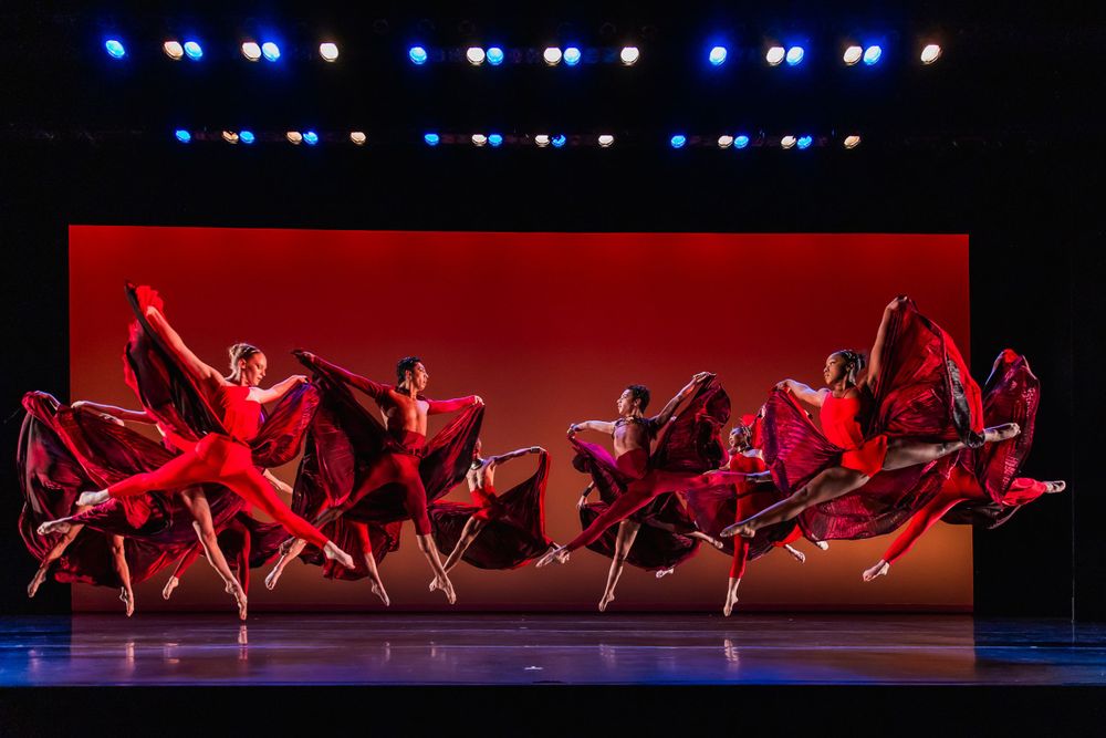 A group of dancers in flowing red costumes perform a synchronized dance sequence on stage.