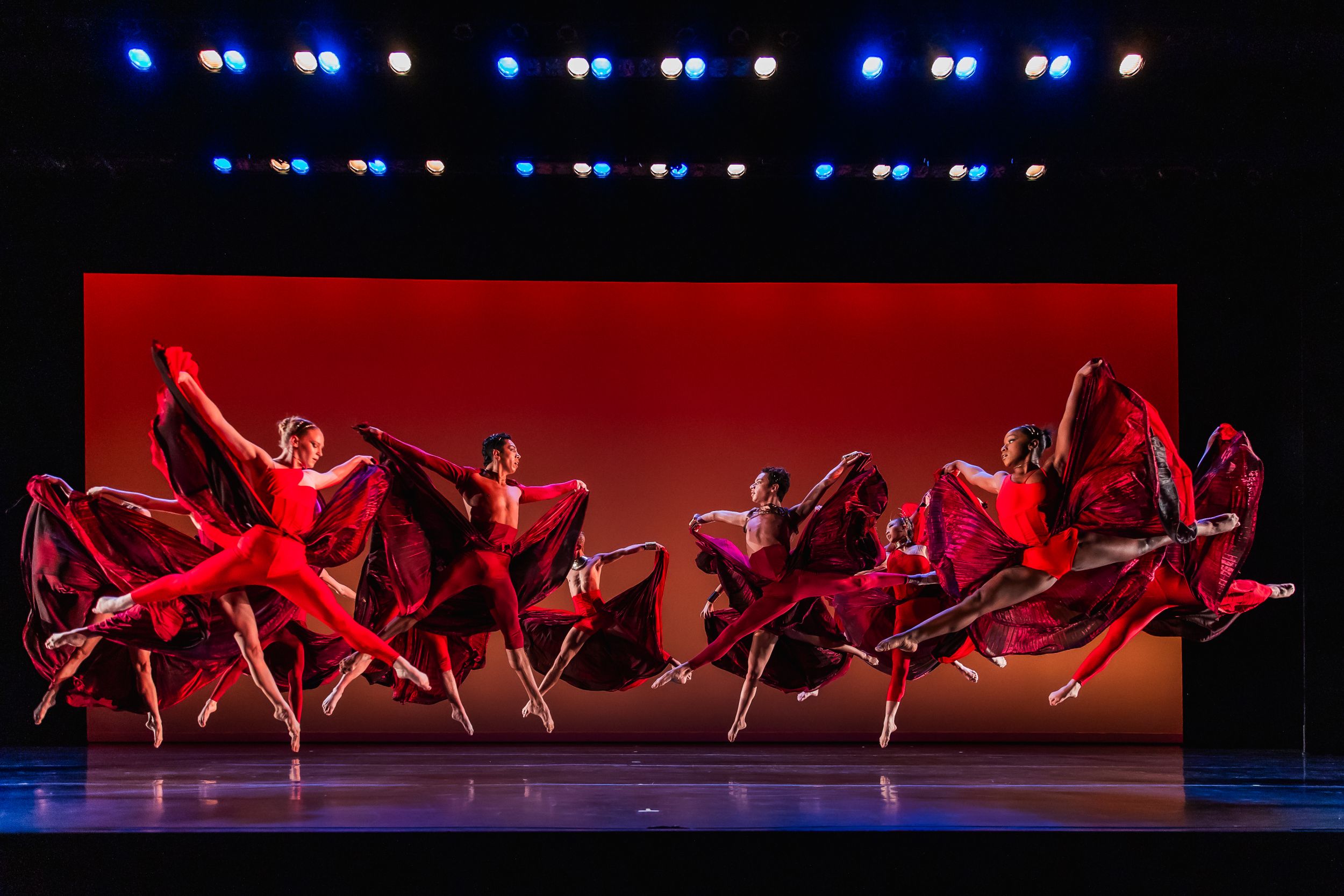 A group of dancers in flowing red costumes perform a synchronized dance sequence on stage.