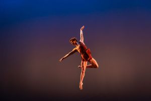 A solo dancer executes a graceful leap in mid-air against a dark blue gradient background.