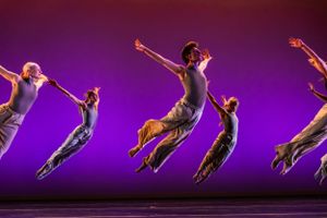 Dancers perform a synchronized leaping sequence against a purple backdrop in a dramatic stage performance.