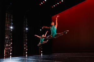 A dancer in teal performs a dramatic split jump against a red-lit backdrop.